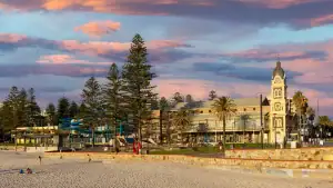 Glenelg Beach at sunset with Norfolk pines, the historic Glenelg Town Hall clock tower, and a playground along the foreshore under a dramatic pink and purple sky